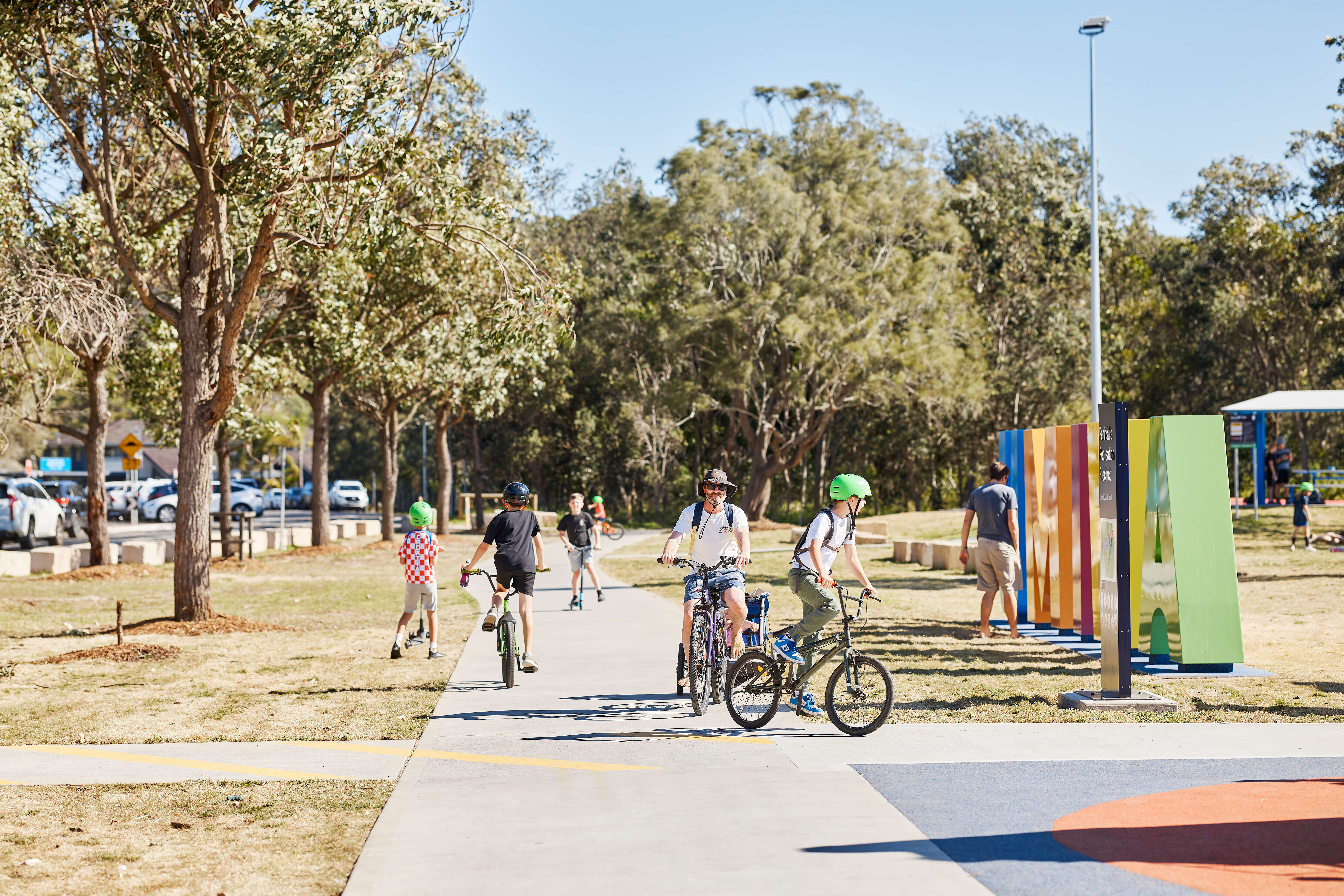 A concrete pathway in a playground with children and adults on bicycles cycling along the path.