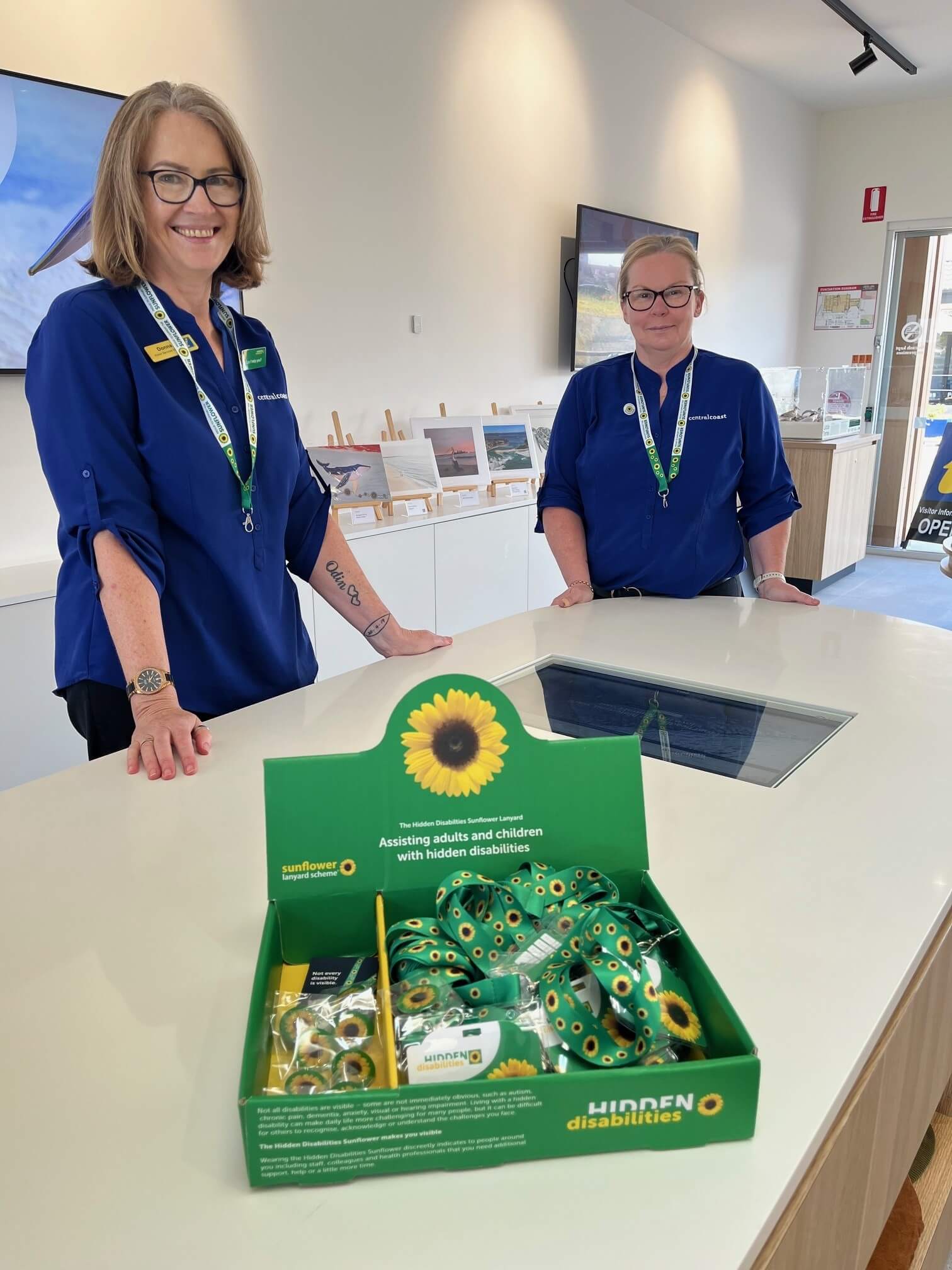 Two women n navy blue shirts with a green merchandise box filled with green lanyards covered in subflowers