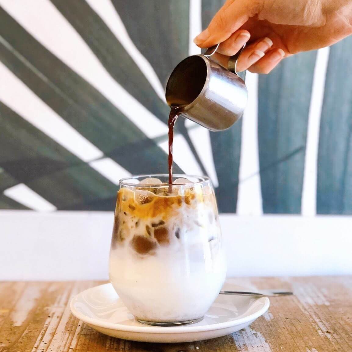 Coffee being poured from a mini silver jug into a glass cup on a saucer filled with milk and ice.