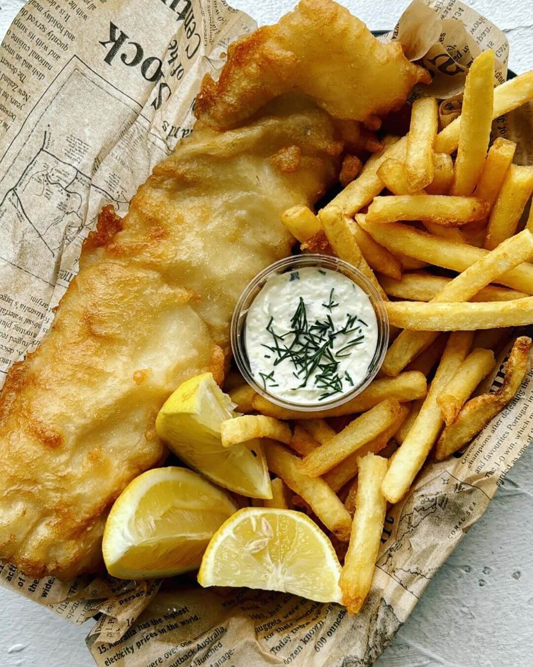 Small takeaway basket filled with battered fish, hot chips, tartare sauce and two slices of lemon