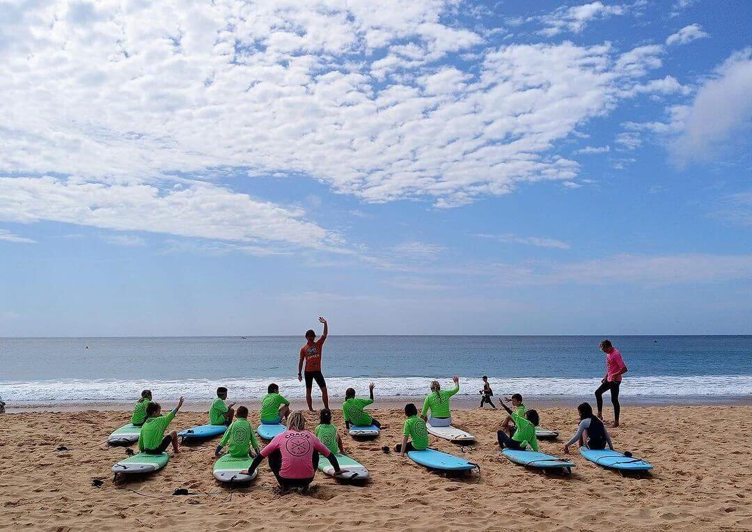 A lively group people learning to surf with colourful surfboards against a sunny beach backdrop.