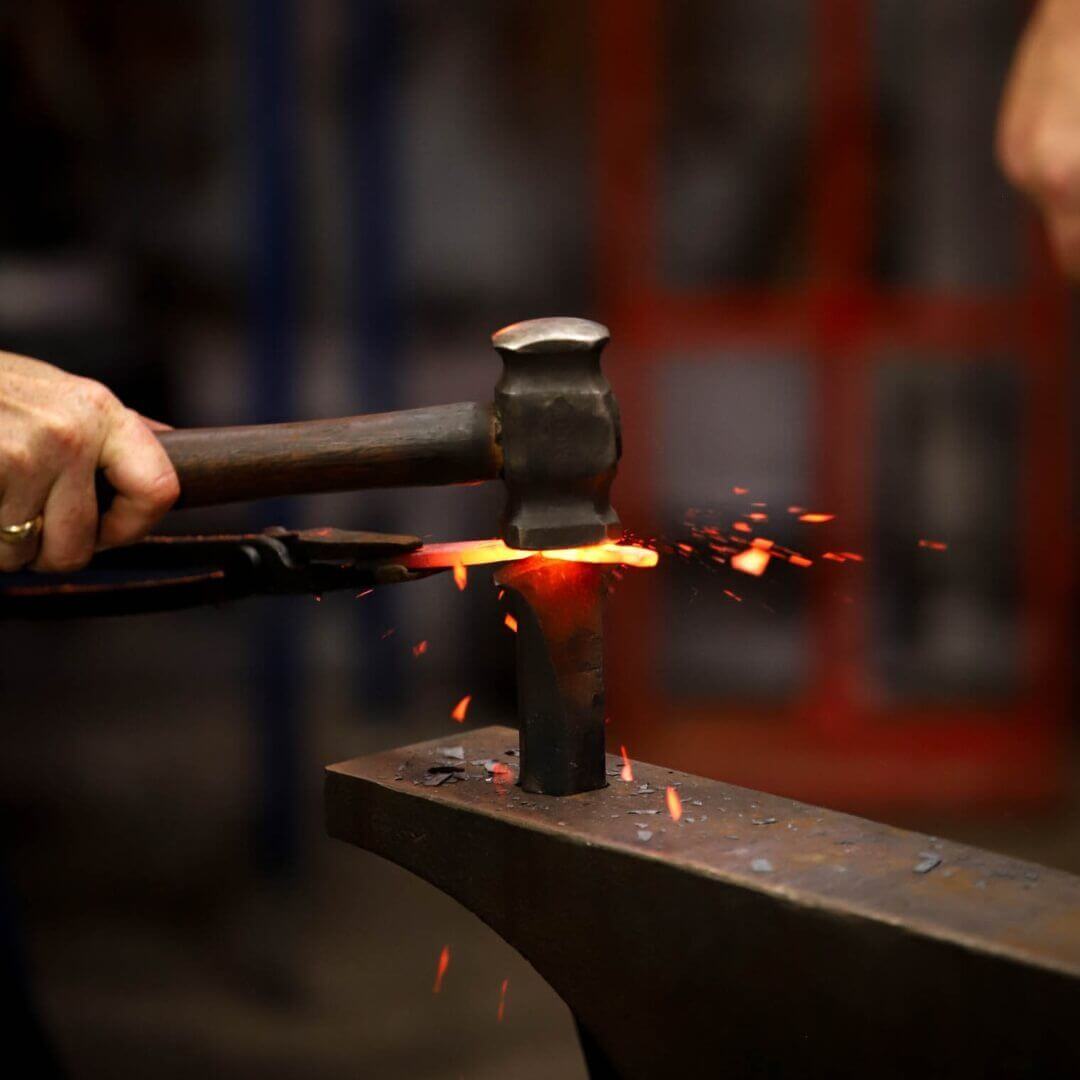 A dark grey metal mallet hitting a glowing, long rectangular piece of metal, ready to be shaped into a bushcraft knife