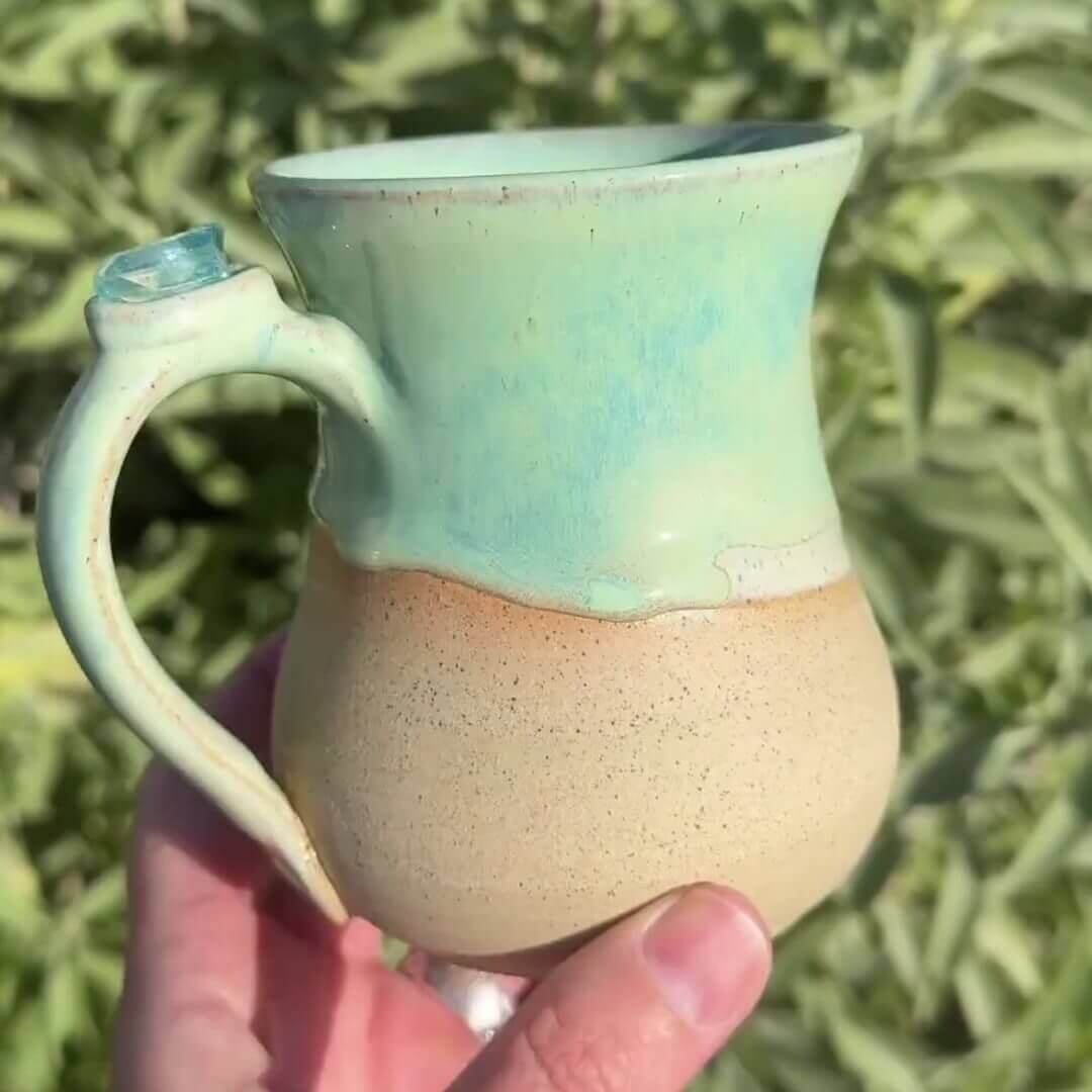 A handmade mug with a sand coloured base. The rim and handle of the mug features a stunning aquamarine glaze and an Aqua Aura gemstone embellished on top of the handle.