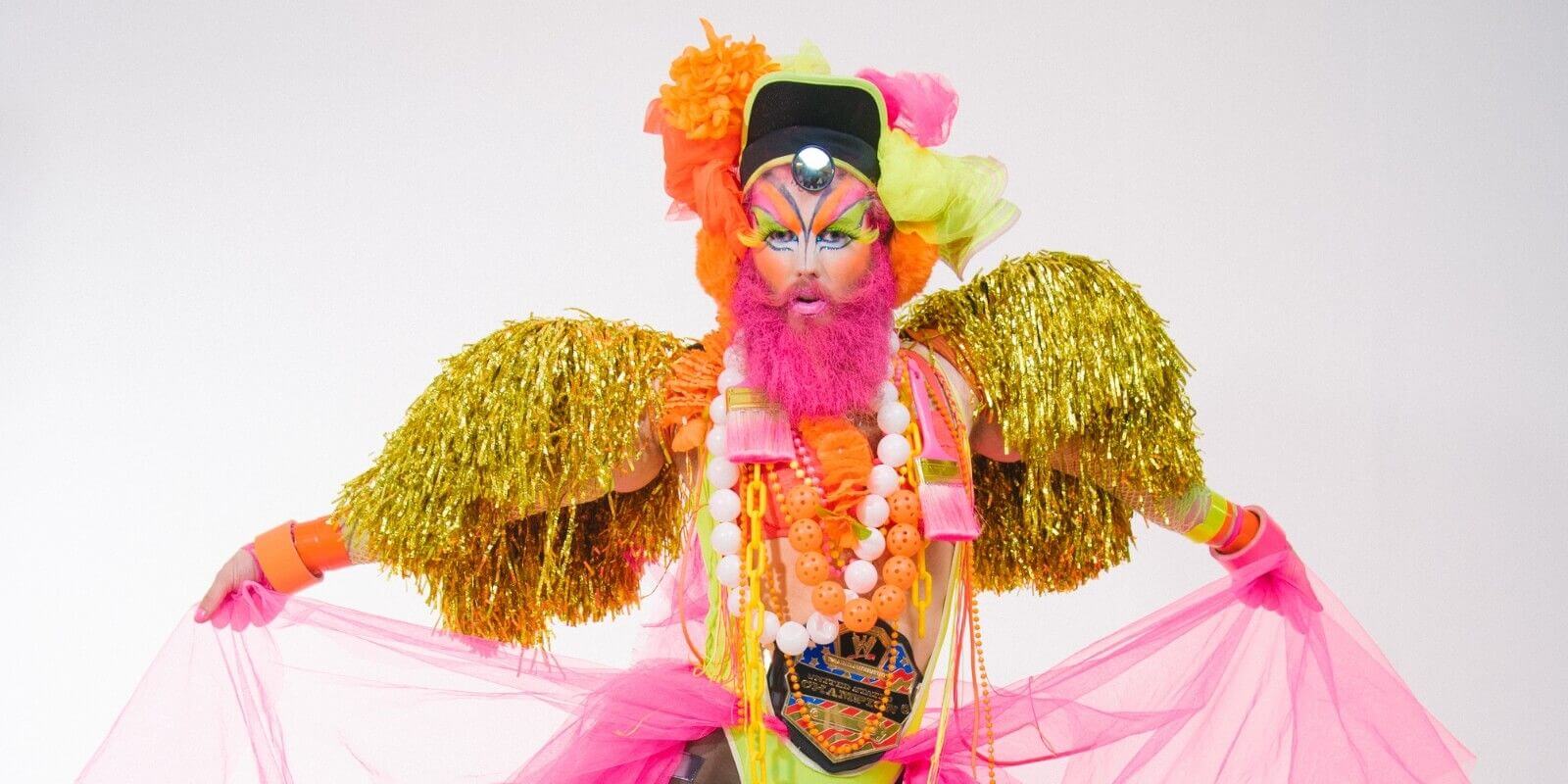 A drag queen with a neon pink beard wearing a neon pink, orange and yellow headpiece and neon dramatic eye makeup