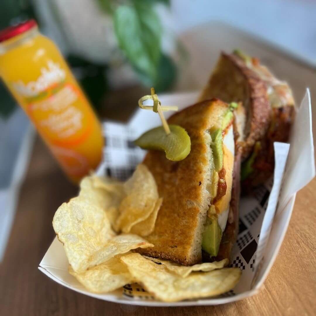 A freshly toasted bacon and egg sandwich, with a side of salty potato crisps and a sweet tangy pickle skewered into the top of the sandwich.