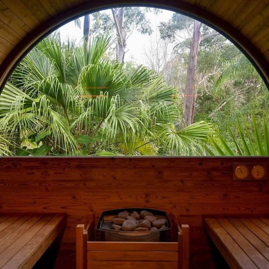 The inside of a round wooden traditional Finnish sauna with two wooden bench seats, the hot rocks in the middle and a semi-circle shaped window looking out onto lush palms.