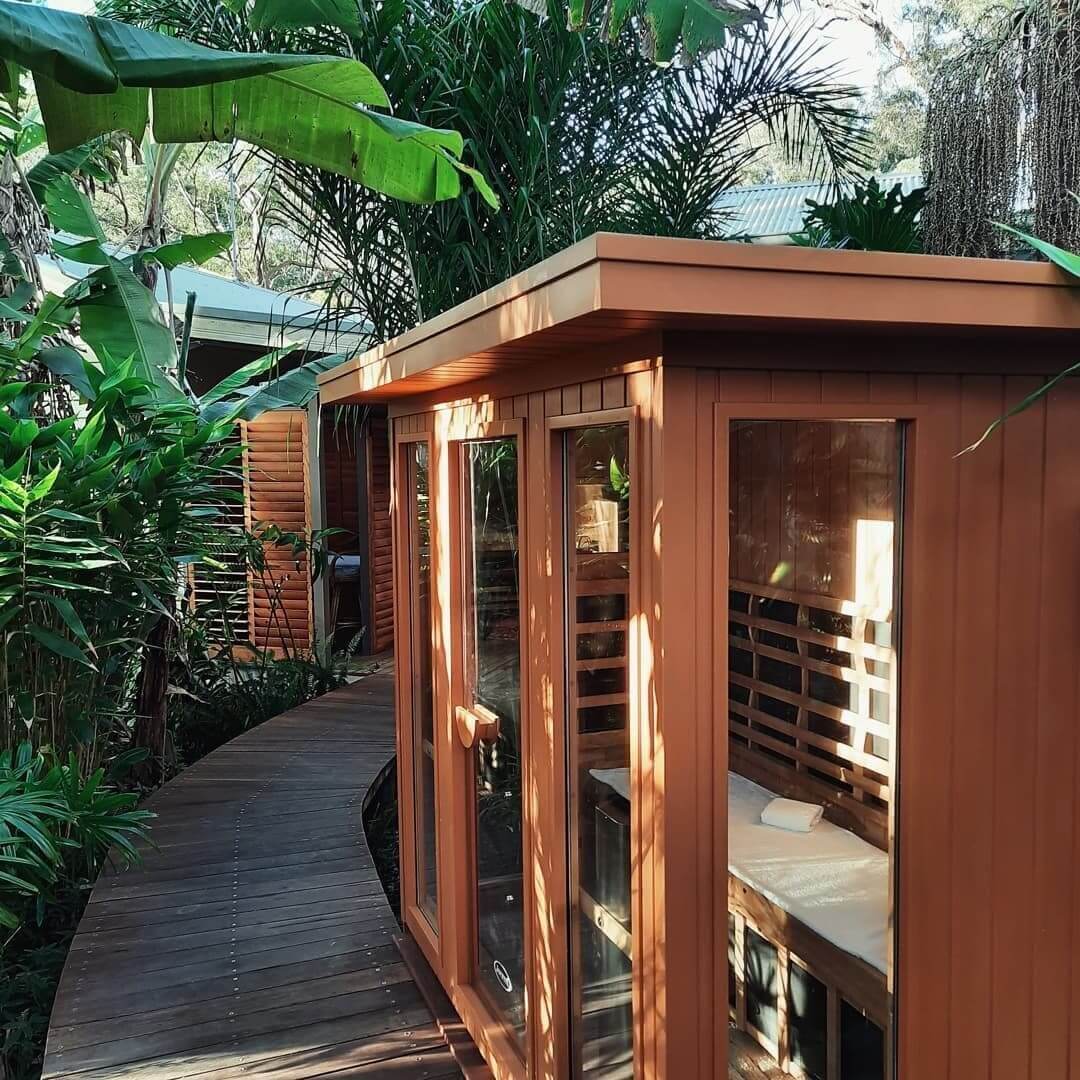 A wooden rectangular infrared sauna sitting beside a wooden boardwalk that is surrounded by lush, tropical foliage.