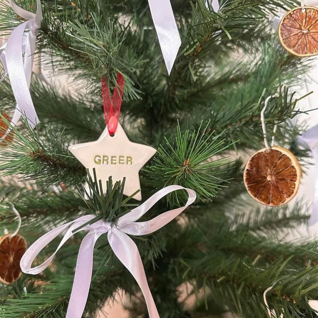 A ceramic star decoration hanging from a Christmas tree, along with dried orange decorations and white ribbon bows tied to the end of each tree branch.