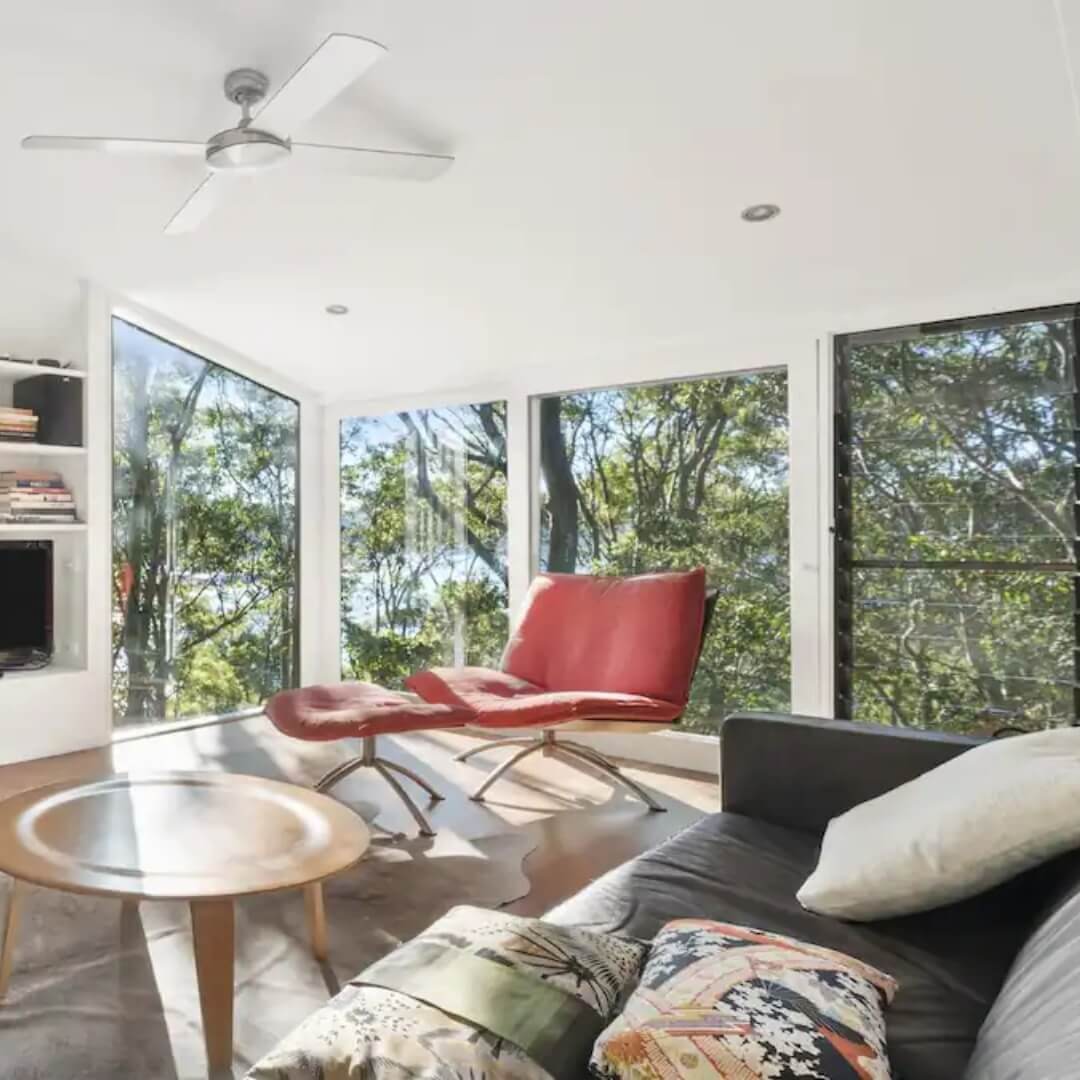 A living room featuring a round wooden coffee table, grey couch, terracotta lounge chair with ottoman. The living room has windows that look out onto Bouddi National Park bushland and the bay.