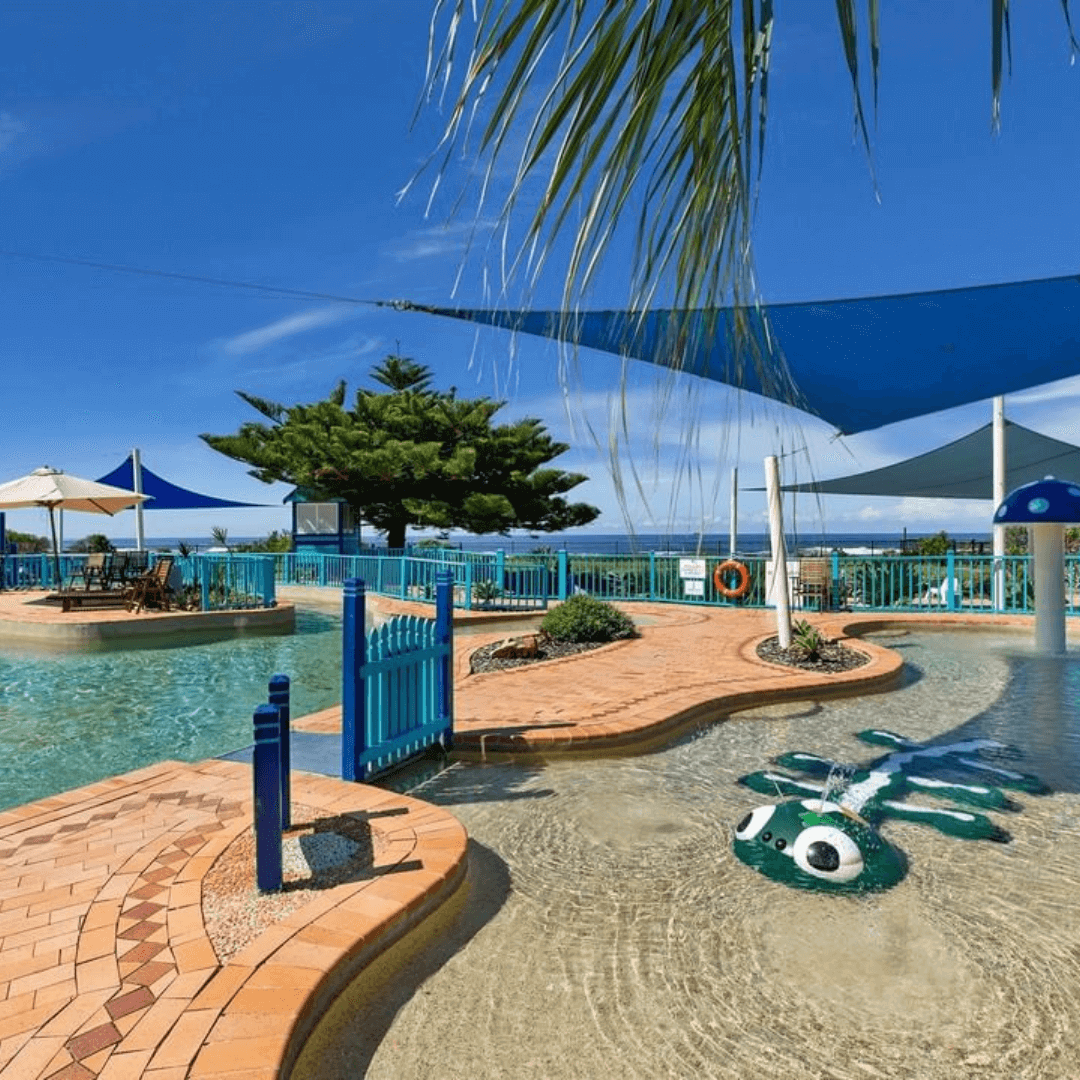 A water park with shallow water. The water park has a mushroom shaped water fountain and on the left there is a deck fitted with beach chairs and cabanas for shade