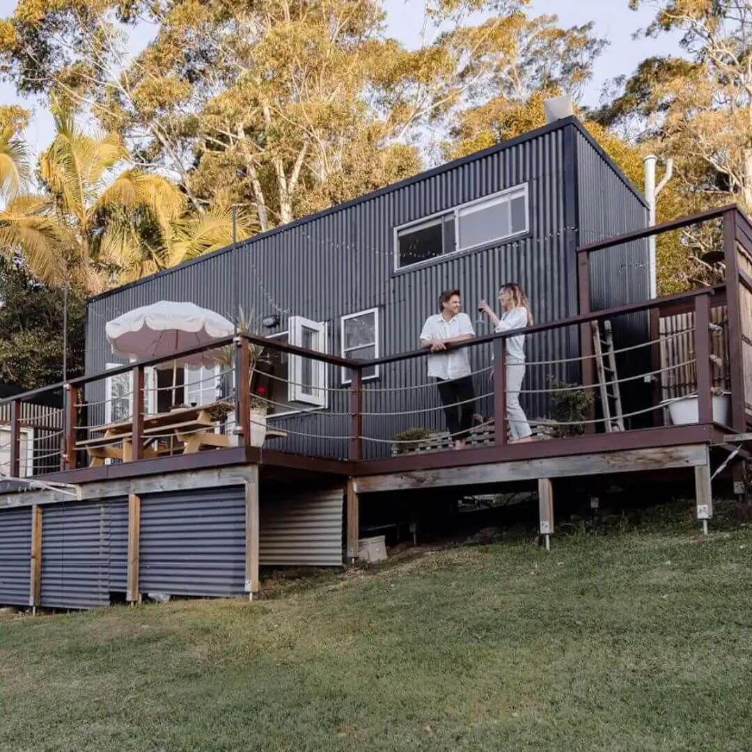 A dark blue-grey tiny house with a couple standing on the deck drinking wine.