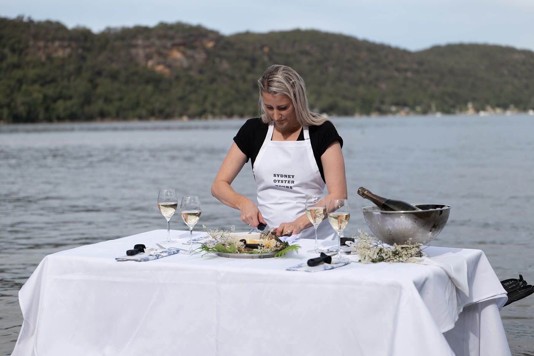 A woman standing at a table in the water, shucking an oyster with a few wine glasses on the table, along with a bottle of wine in an ice bucket