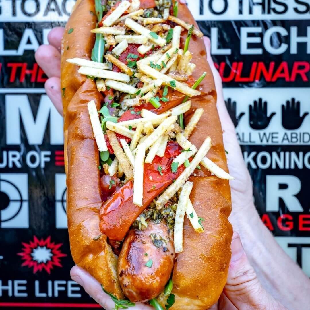 A hot dog bun filled with a chorizo sausage and topped roasted capsicum and small crispy French fries.