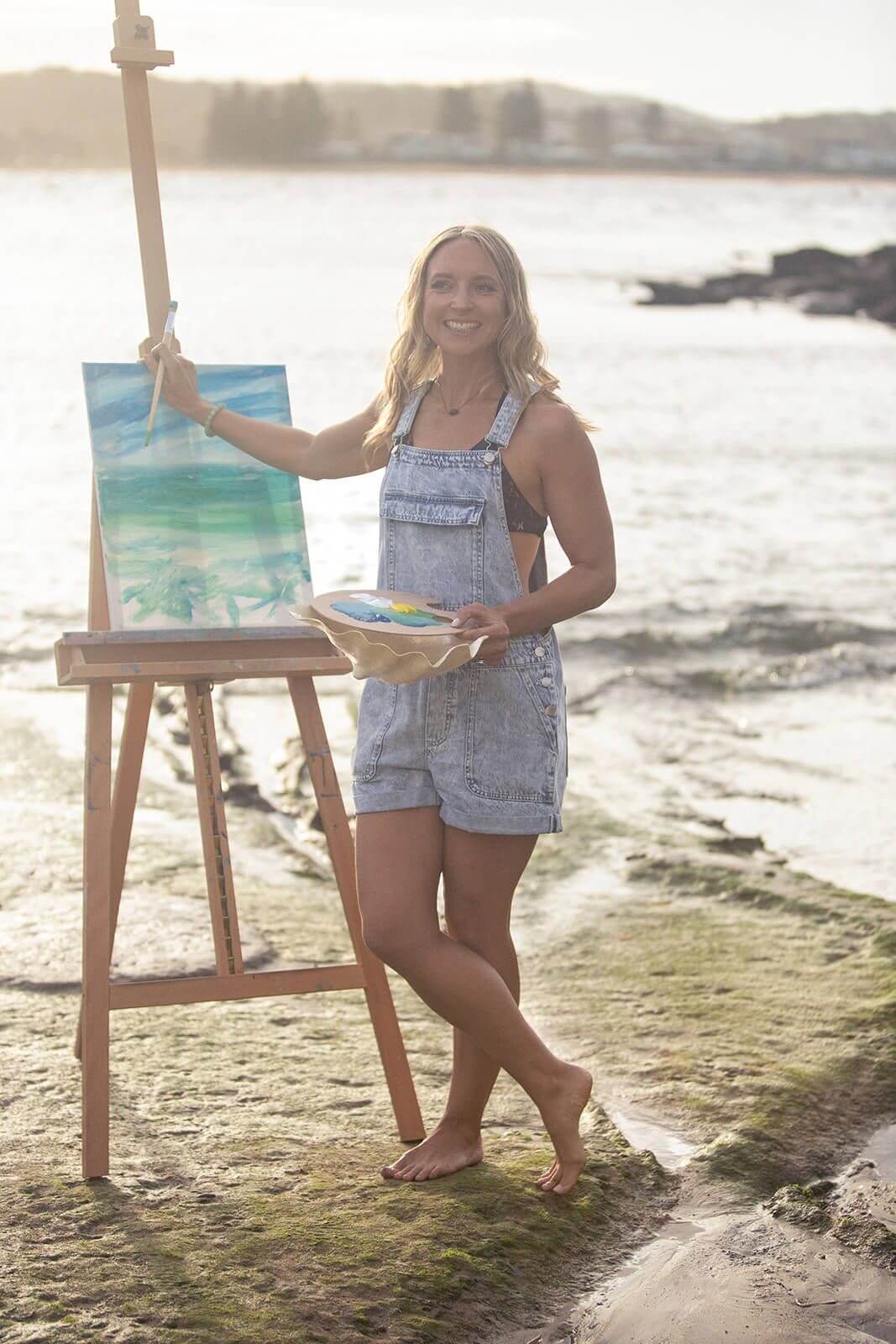 A lady standing on rocks at the beach with a painted canvas