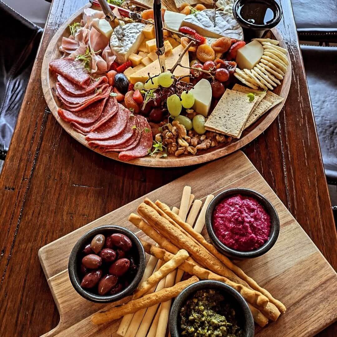 Two charcuterie boards filled with artisan crackers and breadsticks, selection of deli meats and cheeses and fruits and olives.