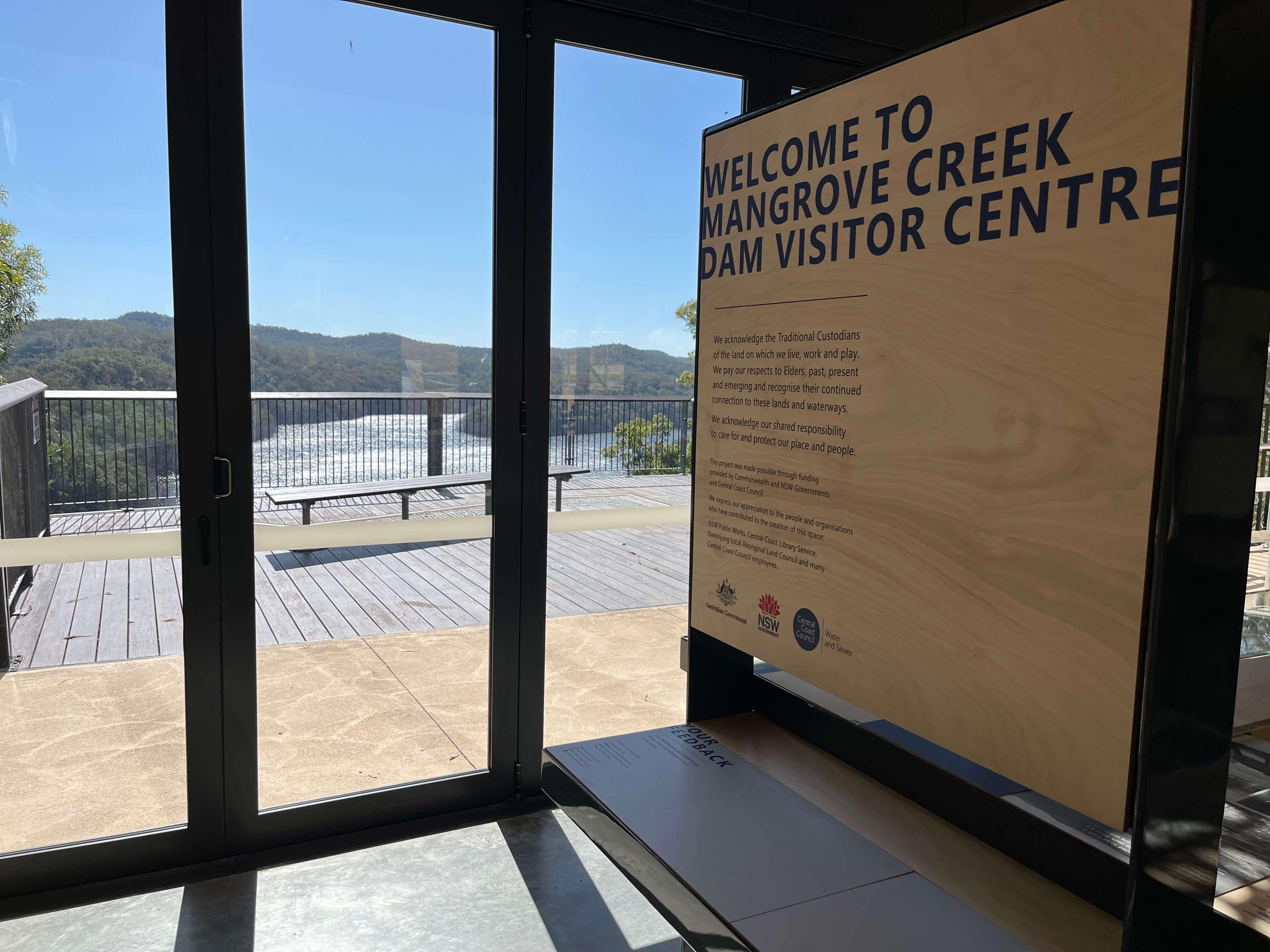 A screen window outlooks to a bushland setting with a wall to the right saying 'Mangrove Creek Dam Visitor Centre'