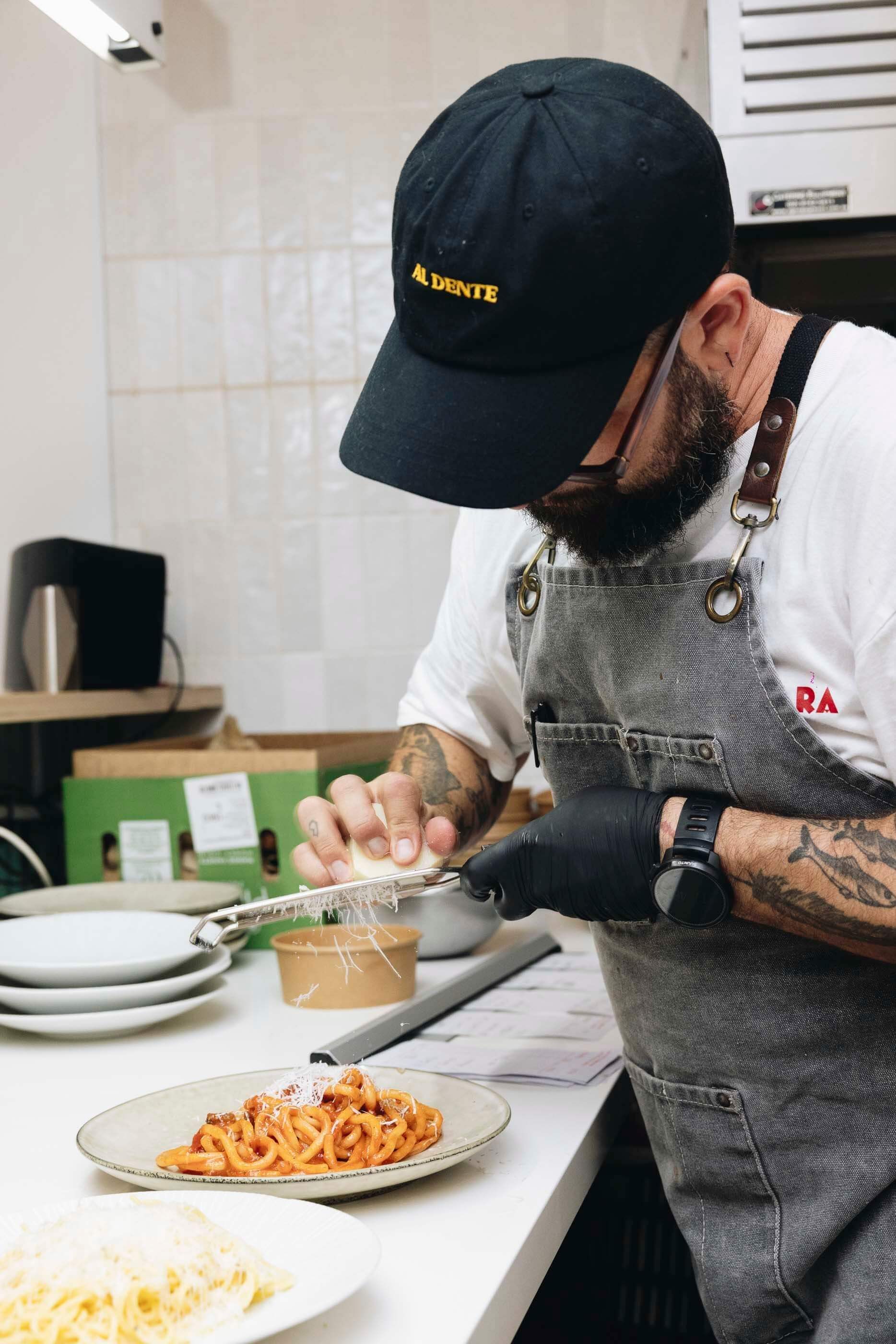 A man wearing a dark blue baseball cap and a grey apron grating cheese over a bowl of pasta