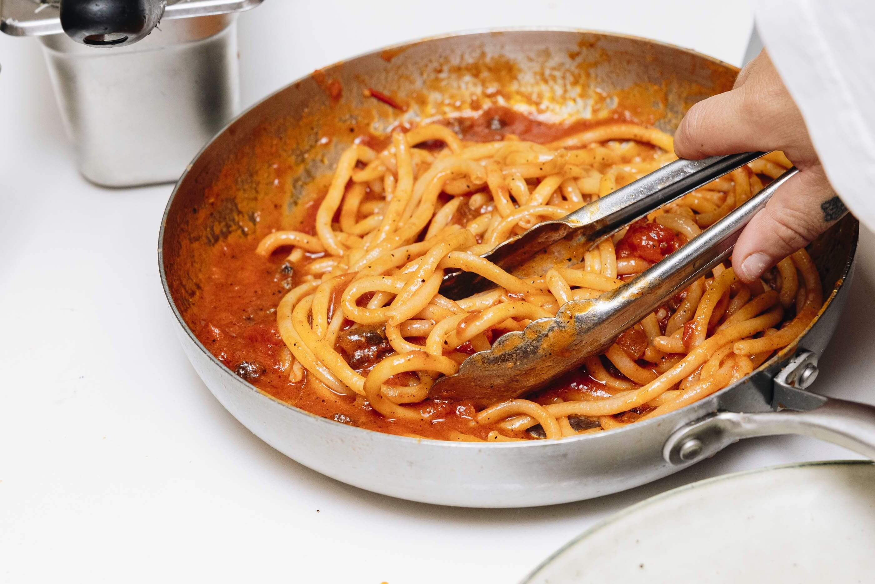 A hand picking up tomato based pasta with tongs from a steel frypan 