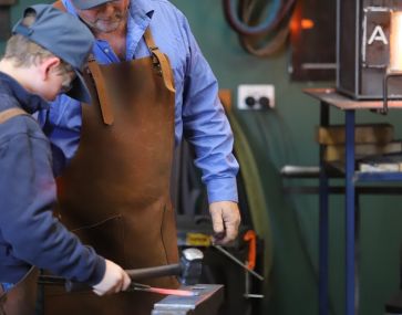 A blacksmith teacher wearing a brown apron overlooking a child hammering a piece of glowing red metal into a long rectangular shape.