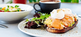 Breakfast serving of eggs benedict, sourdough, avocado, salmon and two poached eggs sprinkled with seasoning and a side of salad. Cappuccino in a black mug with saucer and a stone bowl with fresh food and eggs. 