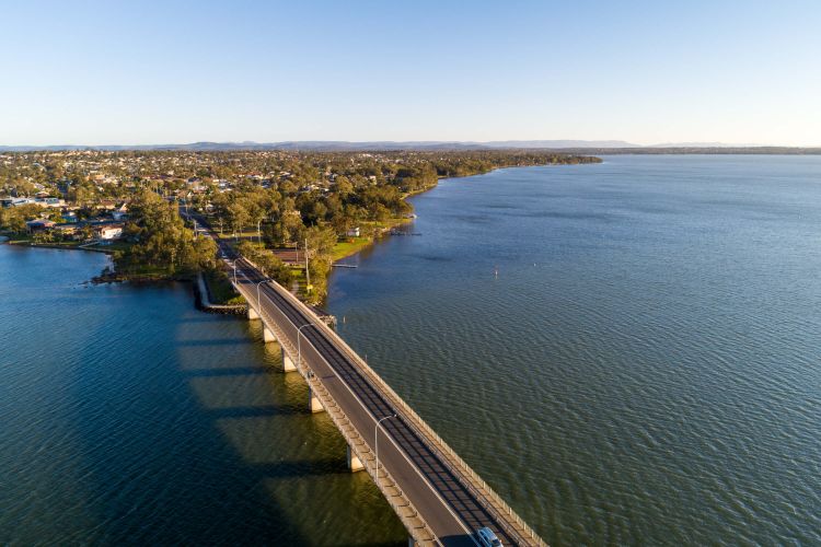 Drone shot of a bridge connecting to a peninsula filled with lush bushland and surrounded by a calm, blue lake.