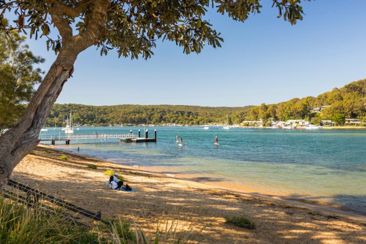 A jetty that juts out into the middle of a calm blue beach. People are paddle boarding in the distance and a couple sits on the sand reading