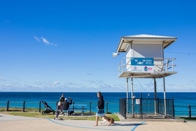 A surf lifesaving tower is on the right and overlooks a bright blue beach. Two men with surfboards walk along the path where the life tower is located.